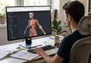 A young man sitting at a desk and using a desktop browser to view an interactive 3D human anatomy model on his monitor. 